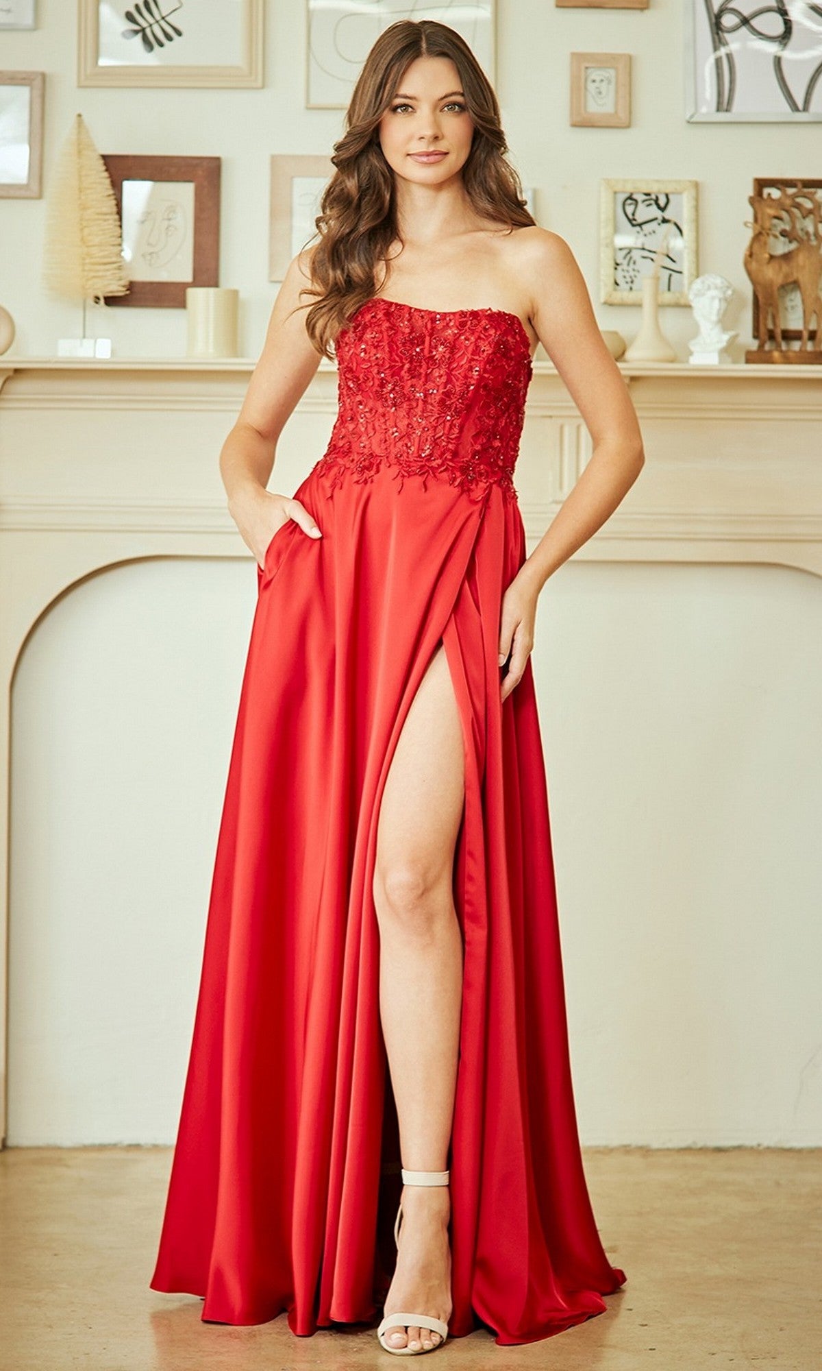 Woman wearing the Amelia Couture 6204 long prom dress, featuring a strapless beaded bodice and thigh-high slit, standing indoors before a decorated mantel.